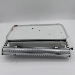 May include: A vintage white metal scale with a rectangular tray and a measurement bar. The scale has a measurement range up to 26 lbs. The scale shows signs of wear and tear, with some rust spots visible on the tray.