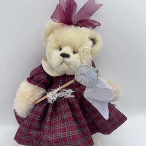 May include: A cream-colored teddy bear wearing a burgundy plaid dress with a white collar and a matching bow. The bear holds a wooden stick with a decorative net and a small, white net.