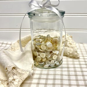 May include: A clear glass jar filled with off-white buttons and a gold-colored safety pin. The jar has a glass lid and is tied with a white ribbon. The jar sits on a checkered beige and white surface.