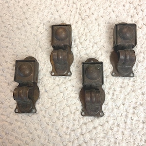May include: Four antique bronze-toned metal cabinet latches. Each latch features a square top with a rounded detail, a curved handle, and a decorative base with screw holes. The latches are arranged on a textured white surface.
