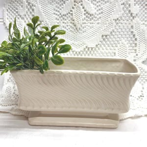 May include: Cream-colored rectangular planter with a textured, wave-like design. The planter sits on a matching base and holds a cluster of green leafy plants, creating a decorative display. A lace doily is in the background.