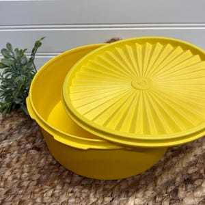 May include: A bright yellow plastic bowl with a matching lid. The lid is partially open, revealing the bowl's interior. The lid features a sunburst design. The bowl is sitting on a woven mat.