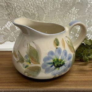May include: Cream-colored ceramic pitcher with a blue floral design. The pitcher has a rounded body, a curved handle, and a spout. The floral design features large blue flowers with green and yellow accents. The rim and handle are trimmed with a light blue.