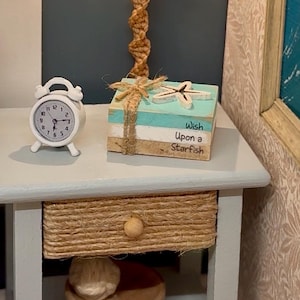 May include: A white alarm clock and a stack of decorative books with the text "Wish Upon a Starfish" sit on a light blue side table. The books are tied with twine and topped with a wooden starfish. The drawer is wrapped in rope.