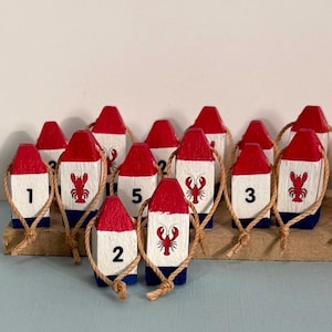 May include: A collection of small wooden lobster buoy ornaments, each painted with a red top, white middle, and blue base. Some feature black numbers, while others display a red lobster illustration. Each buoy has a tan rope loop attached.