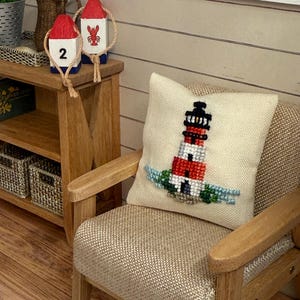 May include: A miniature scene featuring a beige armchair with a lighthouse-themed pillow. Two red and white buoys with rope are on a wooden table. The pillow has a cross-stitch design of a red and white lighthouse with blue accents.