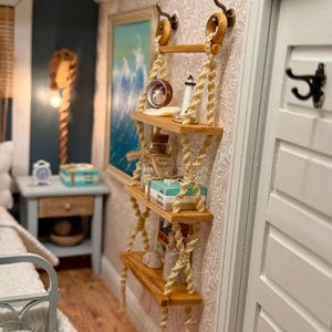 May include: A miniature wooden shelf unit with three shelves, suspended by rope. The shelves hold small books, boxes, and decorative items. The unit is mounted on a patterned wall, with a painting and a door visible in the background.