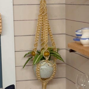 May include: A miniature macrame plant hanger in a light beige color, holding a small ceramic pot with green plants. The hanger is suspended from a wooden ring and mounted on a white wall with horizontal lines. Decorative home decor.