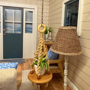 May include: A miniature interior scene featuring a hanging macrame plant holder with a small potted plant. A wooden side table, a floor lamp with a woven shade, and a blue door are also visible. The room has a coastal theme.