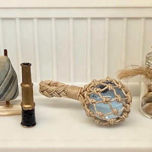 May include: A miniature nautical display featuring a striped stone sail on a wooden base, a small brass-colored telescope, a light blue glass sphere encased in tan woven rope netting, and a clear glass jar filled with small seashells and a cork stopper.