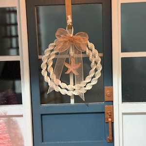 May include: A miniature wreath made of rolled paper, adorned with a peach-colored bow and a starfish pendant, hangs on a blue door. The door has a copper-colored handle and is framed by white trim. The wreath is a decorative accent.