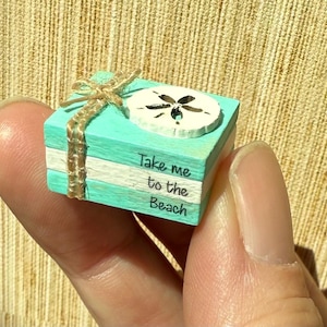 May include: A miniature turquoise and white wooden block with the text "Take me to the Beach". It is decorated with a sand dollar and tied with twine. The block is held in a person's hand.