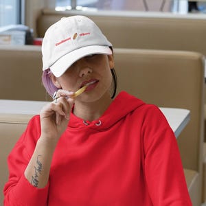 May include: A person wearing a white baseball cap with the text "Condiments Where" in red, and a red hoodie. The person is sitting at a table and eating a french fry. The hoodie has a front pocket.