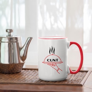 Può includere: Tazza in ceramica bianca con manico e bordo rossi. La tazza presenta una grafica di una mano che tiene un vassoio con la scritta "CUNT". Una teiera d'argento è appoggiata su una superficie di legno.