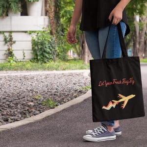 May include: Black tote bag with the text "Let Your Freak Flag Fly" in pink script. A yellow airplane with a rainbow trail is below the text. The bag is held by a person wearing blue jeans and gray sneakers.