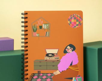 Op de afbeelding: Oranje spiraalgebonden notitieboek met een illustratie van een vrouw in een roze jas en bruine rok. Het ontwerp bevat een platenspeler, een discobal en een plank met planten, allemaal in retrostijl.