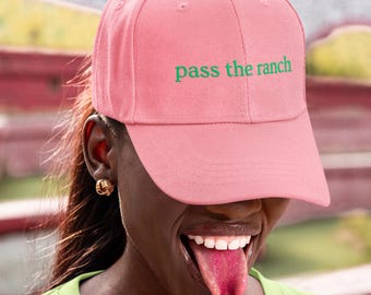 May include: A pink baseball cap with the words "pass the ranch" in green. The cap is worn by a person wearing a green t-shirt. The person is smiling and has their tongue out.