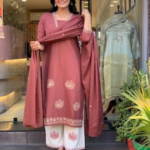 May include: A woman in a mauve salwar kameez. The outfit features a long-sleeved tunic with leaf embroidery, a matching scarf, and white palazzo trousers with leaf patterns. The outfit is displayed in a shop setting.