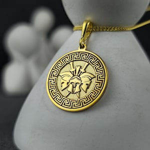 May include: A gold-colored pendant necklace featuring a circular charm with a Greek key border. The charm displays three stylized helmets and a central design. The necklace is displayed on a white, abstract form.