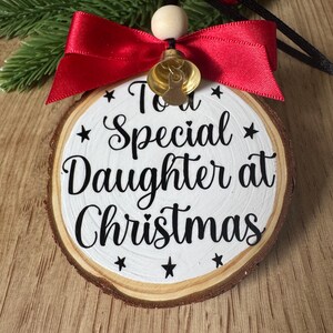 May include: A wooden Christmas ornament with the text "To a Special Daughter at Christmas" in black script. The ornament features a red ribbon, a small gold bell, and a wooden bead. The background includes greenery and red berries.