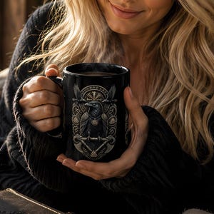 May include: A black ceramic mug featuring a detailed graphic illustration of a raven perched on a branch, surrounded by intricate geometric patterns and symbols. The mug is held by two hands, with steam rising from the top of the dark beverage inside.