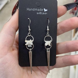May include: Handmade earrings with a distinctive design. Each earring has a silver-coloured metal spike pendant, a small white bone-like charm, and a silver hoop. The black card behind the earrings reads "Handmade with love."