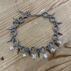 May include: A silver choker necklace featuring metal rings, cone spikes, and small white bone charms. The necklace is displayed on a wooden surface.