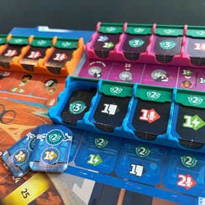 May include: A board game setup featuring various colored plastic trays and game pieces. The trays are orange, pink, and blue, holding cards with numbers and symbols. The game board is visible in the background, with numbered spaces and graphic elements.