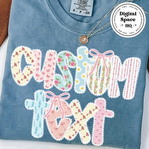 May include: A blue Comfort Colors t-shirt with the words "CUSTOM TEXT" in a patchwork design. The letters are decorated with floral patterns, bows, and stripes. A gold necklace and a brown hat are visible.