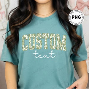 May include: Teal-colored t-shirt with the word "CUSTOM" in a floral design and the word "text" in a cursive font. The shirt is a solid color and has short sleeves.