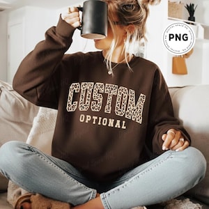May include: A brown sweatshirt with the words "CUSTOM OPTIONAL" in a leopard print design. The sweatshirt is a solid brown color. The image shows a person wearing the sweatshirt.
