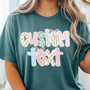 May include: Teal t-shirt with the words "CUSTOM TEXT" in a patchwork design. The letters are decorated with floral, polka dot, and striped patterns, and each letter has a small bow. The shirt is a solid teal color.