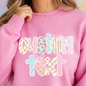 May include: Pink sweatshirt with the words "CUSTOM TEXT" in a floral and striped design. The letters are in various patterns, including roses, daisies, and stripes, with decorative bows. The sweatshirt is a solid pink color.