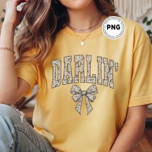 May include: Yellow t-shirt with the word "DARLIN'" in a camouflage pattern and a bow graphic. The shirt is a solid yellow color and has short sleeves. The text and bow are in shades of gray and tan.