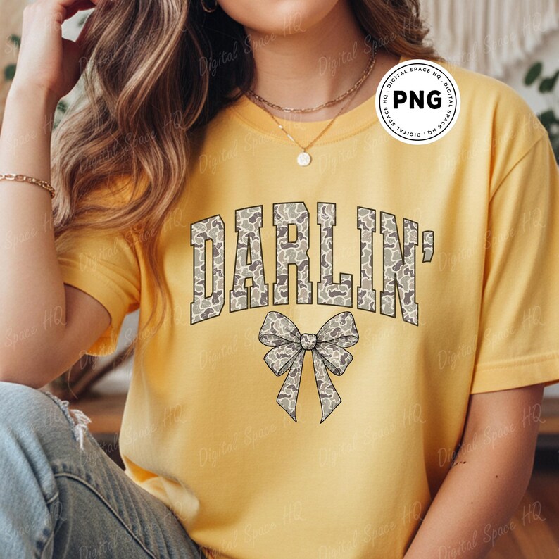 May include: Yellow t-shirt with the word "DARLIN'" in a camouflage pattern and a bow graphic. The shirt is a solid yellow color and has short sleeves. The text and bow are in shades of gray and tan.
