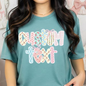 May include: Teal t-shirt with the words "CUSTOM TEXT" in a decorative, floral-patterned design. The letters are adorned with bows and various patterns, including floral prints and stripes. The shirt is a solid teal color.