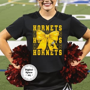 May include: Black v-neck t-shirt with a yellow bow and the word "HORNETS" in yellow block letters. A yellow and black bee graphic is on the bow. The person is holding maroon pom-poms.