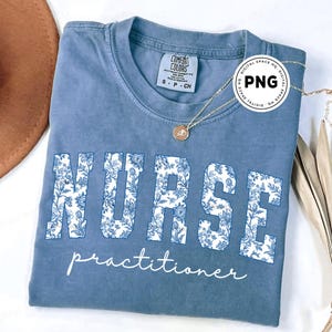 May include: A blue t-shirt with the word "NURSE" in large white floral print letters, and the word "practitioner" in script below. A gold necklace with a pendant is draped on the shirt.