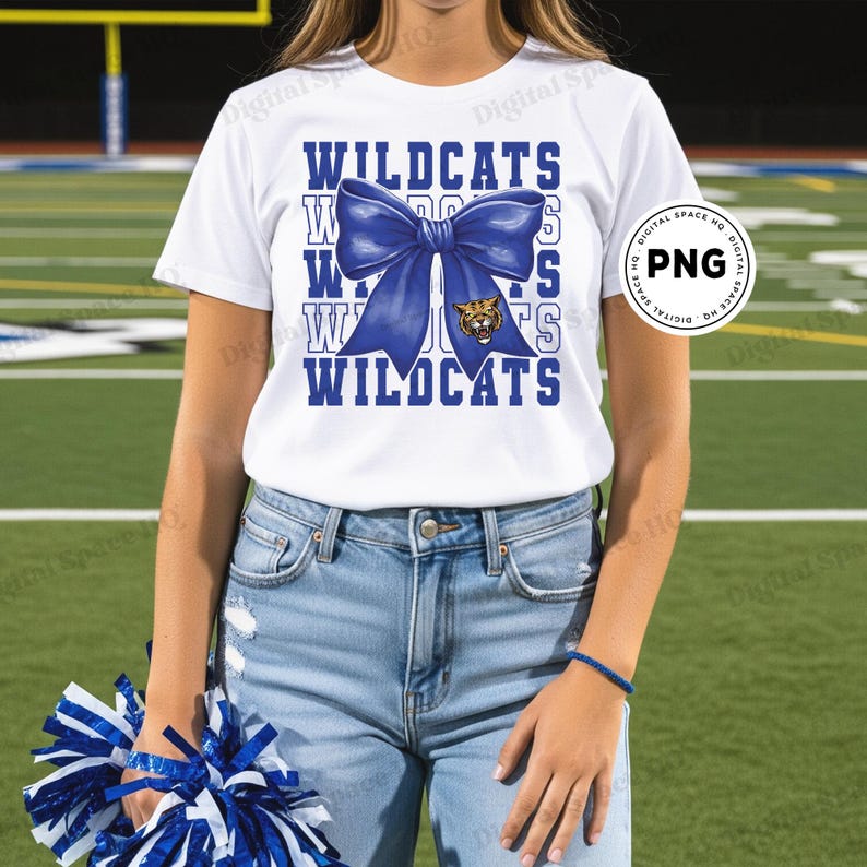 May include: White t-shirt with the word "WILDCATS" in blue block letters, a blue bow graphic, and a tiger mascot. The shirt is paired with blue jeans. A person is holding blue and white pom-poms.
