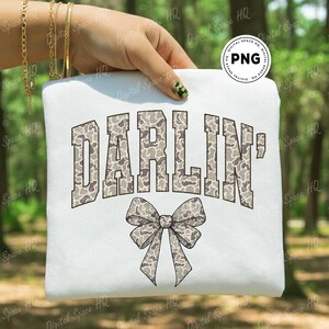 May include: White sweatshirt featuring the word "DARLIN'" in a camouflage pattern, arched above a matching bow graphic. The design is in shades of gray, brown, and white. The sweatshirt is held up by a hand.