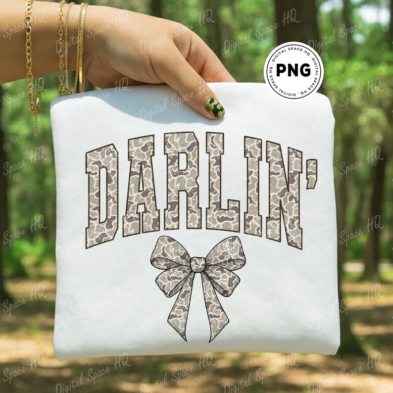 May include: White sweatshirt featuring the word "DARLIN'" in a camouflage pattern, arched above a matching bow graphic. The design is in shades of gray, brown, and white. The sweatshirt is held up by a hand.