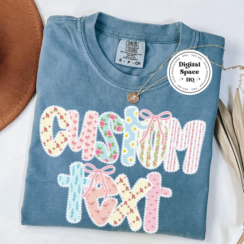 May include: A blue Comfort Colors t-shirt with the words "CUSTOM TEXT" in a patchwork design. The letters are decorated with floral patterns, bows, and stripes. A gold necklace and a brown hat are visible.