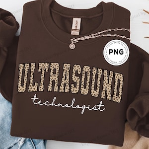 Puede incluir: Una sudadera marrón con la palabra "ULTRASOUND" en letras con estampado de leopardo sobre la palabra "technologist" en escritura blanca. Se ve un collar plateado con un colgante.
