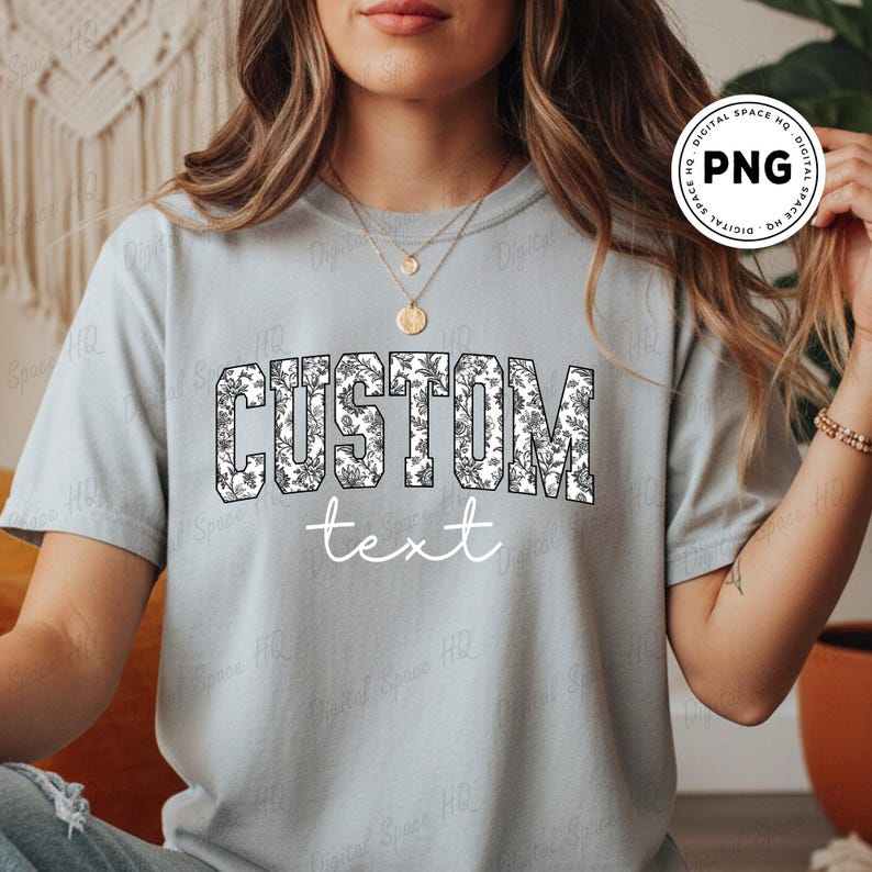 May include: A light gray t-shirt with the word "CUSTOM" in large, bold, black letters filled with a white floral pattern. Below, the word "text" is written in a simple, white font. The shirt is a casual style.