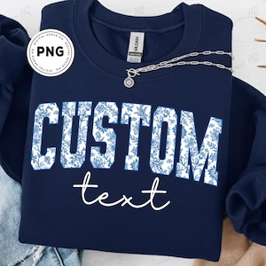 May include: Navy blue sweatshirt with the word "CUSTOM" in large, white, floral-patterned letters. Below, the word "text" is written in a cursive font. A silver necklace with a pendant is draped on the sweatshirt.