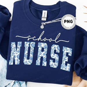 May include: A navy blue sweatshirt with the words "school NURSE" in white script and floral print. The sweatshirt has a crew neck and long sleeves. A silver necklace is draped across the top of the sweatshirt.