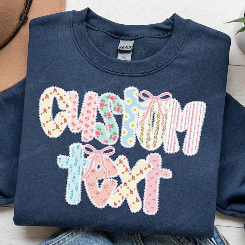 May include: Navy blue sweatshirt with the words "CUSTOM TEXT" in a floral and striped design. The letters are outlined in white and adorned with pink bows. The sweatshirt is a comfortable, casual garment.