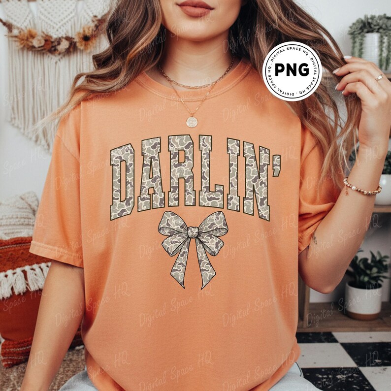 May include: Orange short-sleeved t-shirt with the word "DARLIN'" in a camouflage pattern across the chest. A matching camouflage bow is centered below the word. The shirt is a solid orange color.