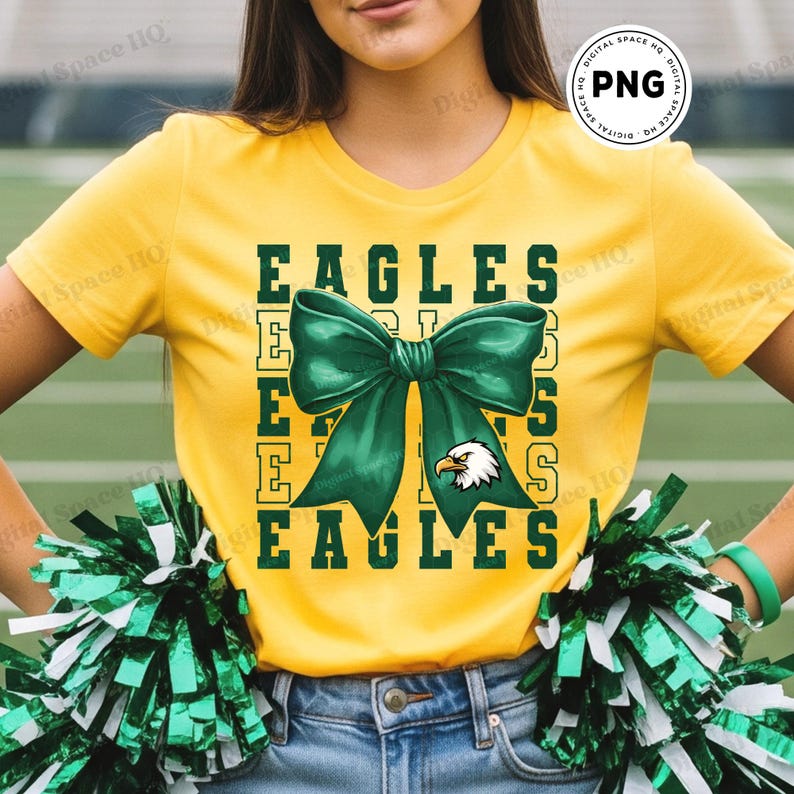 May include: A yellow t-shirt with the word "EAGLES" in green block letters. A large green bow graphic is centered on the shirt, with an eagle head emblem. The person is wearing blue jeans and holding green and white pom-poms.
