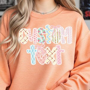 May include: Peach-colored sweatshirt with the words "CUSTOM TEXT" in a patchwork design. The letters are decorated with floral patterns, bows, and stripes. The sweatshirt is a casual, comfortable style.